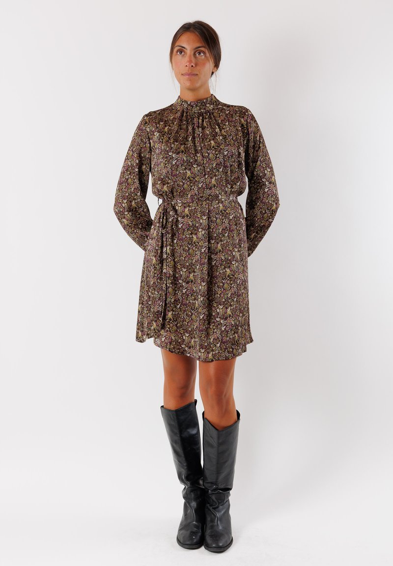 Abito floreale in tonalità scure con un collo alto e maniche lunghe. Presenta una vita con lacci e una gonna fluida. Indossato con stivali neri fino al ginocchio.