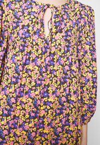 Långärmad blommig klänning med knytkrage. Har rosa, gula och blå blommor på en brun bakgrund. Lättviktsmaterial med rynkade ärmar.