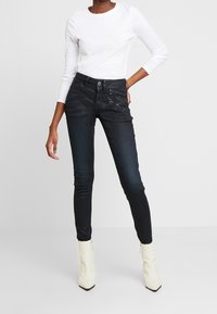 Svarta smala jeans med dragkedjeslut i framsidan, hög midja och en slät yta, kombinerade med en åtsittande vit långärmad skjorta och krämfärgade ankelstövlar.