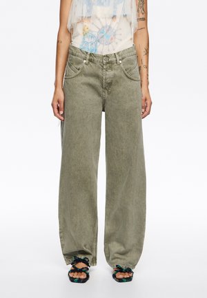 Helle olivgrüne Schlagjeans mit frontalen Taschen und hoher Taille, aus weichem Denim. Begleitet von strukturierten, mehrfarbigen Sandalen.