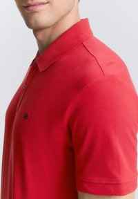 Rød polo skjorte lavet af bomuld, med en klassisk krave, to knapper og et lille logo på brystet. Glat tekstur og tætsiddende design.