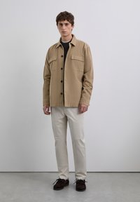 Joven de pie con una chaqueta de camisa color arena, suéter oscuro, pantalones beige claros y zapatos marrón oscuro, contra un fondo gris liso.