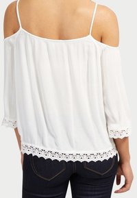 Biała bluzka z odkrytymi ramionami, wykonana z fakturowanego materiału, z rękawami do połowy przedramienia i koronkową lamówką na dole. Noszona z ciemnymi dżinsami.