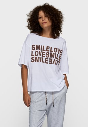 Biała, oversize koszulka z brązowym napisem "SMILELOVE LOVESMILE" w różnych orientacjach; połączona z pasiastymi spodniami od piżamy.