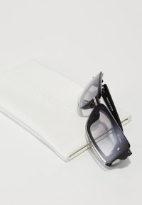 Mustat aurinkolasit suorakulmaisen muodon ja sileän pinnan kanssa, esillä valkoisella nahkapussilla, jossa on välillä "Le Specs."