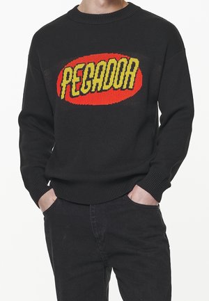 Schwarzer Strickpullover mit einem roten Oval und fettgedrucktem gelbem Text "PEGADOR", getragen von einer Person mit den Händen in den Taschen einer schwarzen Hose.