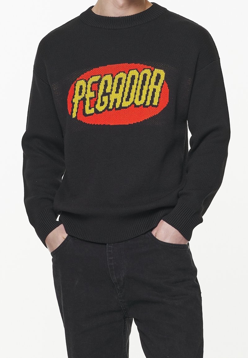 Pull en tricot noir avec un ovale rouge et un texte jaune vif "PEGADOR" porté par une personne avec les mains dans les poches d'un pantalon noir.