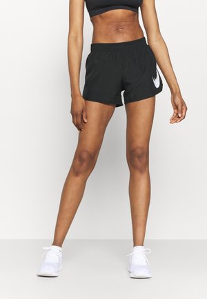 Athletische Person, die schwarze Nike-Shorts, einen schwarzen Sport-BH und weiße Sneakers trägt und mit auseinanderstehenden Beinen vor weißem Hintergrund steht.