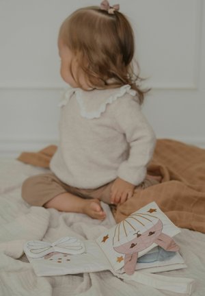 Väikelaps istub pehmetel pleedidel, seljas beež riietus valge kraega, vaatab küljele, kõrval avatud kangaraamat loomapiltidega.