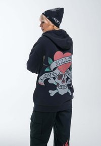 Felpa nera con un design di teschio colorato con un cuore, accompagnata da un banner con la scritta "L'AMORE UCCIDE LENTAMENTE." I dettagli di accento includono foglie e ossa incrociate.