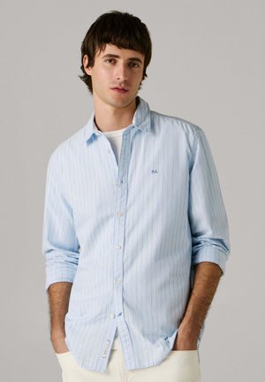 Noor mees tumedate juustega, kellel seljas hele sinine, triibuline, pika varrukaga nööbitav särk valge särgi peal, käed taskutes.