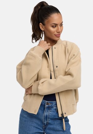 Beige bolyhos bomber dzseki bordás mandzsettával és szegéllyel, elöl cipzárral és patent gombokkal, sötét farmernadrág felett viselve.