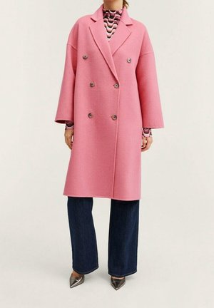 Femme portant un long manteau rose croisé sur un haut à motifs, un jean large foncé et des chaussures pointues métalliques, debout.