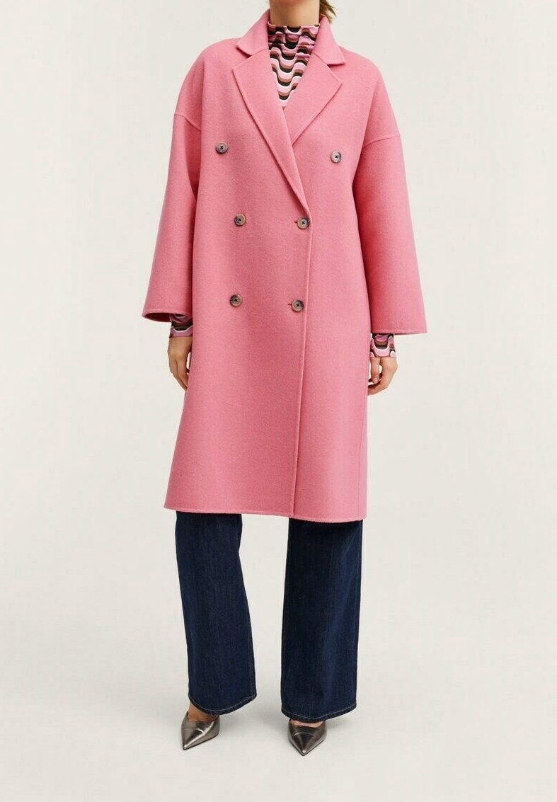 Femme portant un long manteau rose croisé sur un haut à motifs, un jean large foncé et des chaussures pointues métalliques, debout.