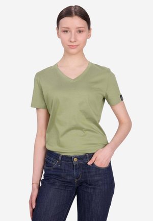 Junge Frau mit dunklem Haar trägt ein hellgrünes T-Shirt mit V-Ausschnitt und dunkelblaue Jeans, steht mit einer Hand in der Tasche vor einem einfarbigen Hintergrund.