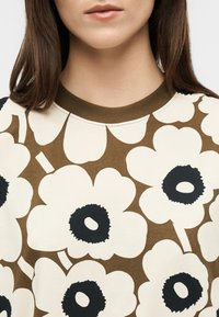 Γυναίκα που φοράει καφέ και κρεμ μπλούζα με μεγάλα κρεμ λουλούδια με μαύρα κέντρα, καφέ γιακά, με ίσια καστανά μαλλιά, εμφανιζόμενη από το πηγούνι και κάτω.