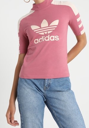 Kobieta ubrana w różową bluzkę z golfem marki Adidas z białymi paskami na rękawach i jasnoniebieskie jeansy, stojąca z jedną ręką na biodrze.