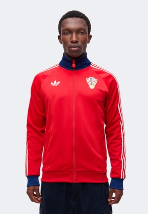 Giovane uomo che indossa una giacca rossa Adidas con zip, colletto e polsini blu, strisce bianche sulle maniche e stemma di squadra sportiva sul petto sinistro.