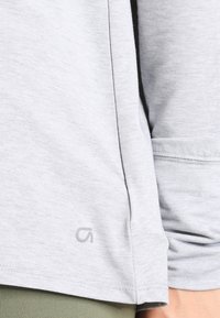 Helles graues Langarmhemd aus weichem Stoff; mit dezentem Branding am Saum und Details an den Bündchen. Glatte Textur und lässige Passform.