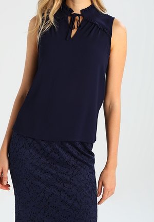 Vrouw die een mouwloze marineblauwe blouse met een striksluiting draagt en een marineblauwe kanten rok, staand tegen een effen achtergrond.