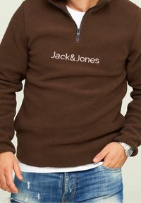 Sudadera de forro marrón con cuello de media cremallera, que presenta el logo "Jack&Jones" en blanco. Textura suave y corte relajado, combinada con vaqueros.