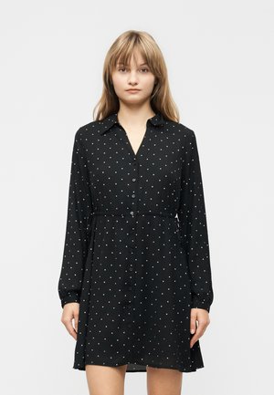 Jeune femme portant une robe noire à manches longues avec des boutons et des pois blancs, debout devant un fond clair uni.