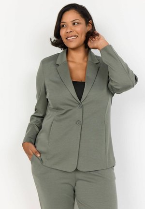 Blazer verde oliva con due bottoni, revers a punta e maniche lunghe. Tessuto liscio, vestibilità su misura, abbinato a un top nero.