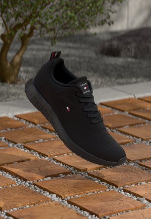 Scarpa atletica nera con tomaia in tessuto testurizzato, lacci piatti e suola in gomma. Presenta un dettaglio del logo e un design minimalista. Posizionata sopra un sentiero in pietra.