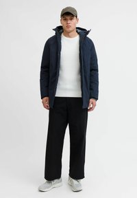 Marineblaue isolierte Jacke mit Reißverschluss, kombiniert mit einem weißen Rippstrickpullover, getragen mit schwarzen weiten Hosen und grauen Sneakern.