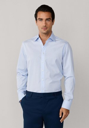 Homme portant une chemise à manches longues à rayures bleu clair, rentrée dans un pantalon bleu foncé, debout les mains dans les poches.