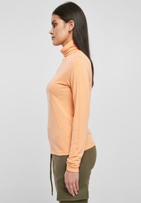 Top a maniche lunghe pesca con una texture liscia, design aderente e colletto leggermente alto, abbinato a pantaloni verdi con cordoncino.