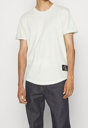 Hellgrünes Baumwoll-T-Shirt mit kurzen Ärmeln und rundem Ausschnitt, mit einem schwarzen Logotag am Saum. Kombiniert mit dunkelblauen Denim-Hosen.