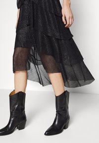 Svart lagerklänning i genomskinligt tyg, med vågig textur. Bärs med spetsiga svarta läderstövlar med blockklack.