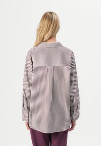 Vrouw met lang blond haar die een losse witte shirt met bruine verticale en horizontale strepen en paarse broek draagt, met de rug naar de camera gekeerd.