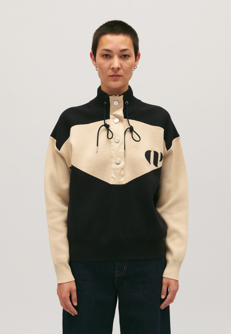 Pull-over en blocs de couleur avec des panneaux noirs et beige, un col montant, une fermeture à boutons-pression à l'avant, des cordons de serrage et un logo brodé sur la poitrine.