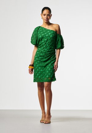 Vrouw draagt een groene one-shoulder jurk met pofmouw, houten armbanden en beige open sandalen, staand voor een effen achtergrond.