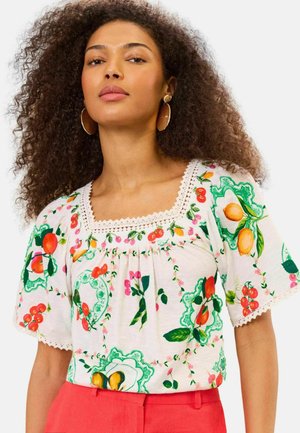 Mujer con cabello rizado que lleva grandes pendientes redondos, una blusa blanca con estampado floral y bordes festoneados, y pantalones rojos de cintura alta.