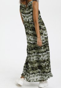 Maxi jurk in groen en zwart abstract patroon, mouwloos ontwerp, vloeiende stof met geplooide taille, gecombineerd met witte plateau schoenen.