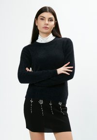 Dunkelblaues, flauschiges Sweatshirt mit langen Ärmeln, kombiniert mit einem schwarzen Rock, der dekorative silberne Blumenverzierungen aufweist.