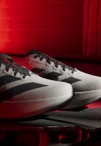 Weiße Laufschuhe mit schwarzen Streifen und Schnürsenkeln, die auf einer dunklen Oberfläche vor einem rot beleuchteten Hintergrund liegen.