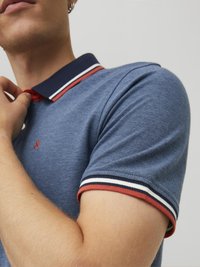 Sininen neulottu polo paita, jossa on tumma kaulus ja punaiset yksityiskohdat. Huomattavat raidalliset hihansuut punaisena, valkoisena ja laivaston sinisenä. Lähikuva kauluksen alueesta.