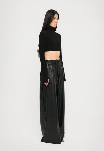 Sieviete, tērpta melnā īsajā turtleneck krekliņā un platajās melnās ādas biksēs, stāv pretī vienkāršai gaišai fona.