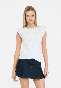 Top bianco a maniche corte con arricciature sul davanti e un collo decorativo con perline, abbinato a una gonna in denim scuro con pieghe.