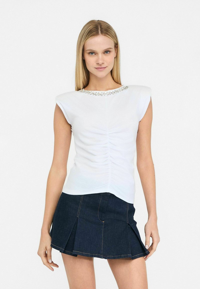 Top bianco a maniche corte con arricciature sul davanti e un collo decorativo con perline, abbinato a una gonna in denim scuro con pieghe.