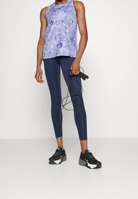 Débardeur violet à motifs associé à des leggings bleu marine et des chaussures de sport noires. Le modèle tient une corde à sauter noire. Textures lisses, coupe ajustée.