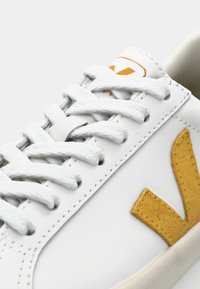 Valge nahkne tennisepaar kollase suede logoga, valged sidemed ja beež tald. Mootori detailid keele ja külgede ääres.