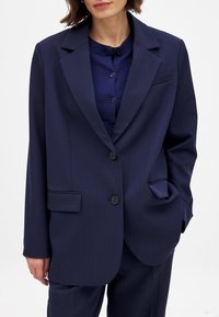 Navy blazer made of smooth fabric, featuring a notched lapel, two front pockets, and two buttons. Worn over a dark blue top.