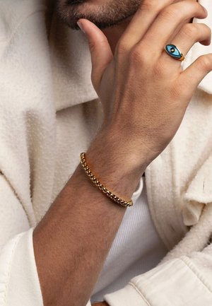 Händ eines Mannes in der Nähe des Gesichts, der ein goldfarbenes Perlenarmband und einen blauen bösen Blick-Ring am Ringfinger trägt, gekleidet in einem cremafarbenen Mantel und weißen Hemd.