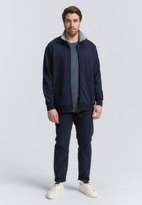 Giacca a zip blu marina con colletto grigio chiaro, abbinata a una t-shirt grigia, pantaloni scuri e sneakers bianche. Tessuto morbido e vestibilità rilassata.