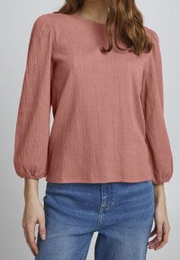 Blusa rosa texturizada con mangas largas abullonadas y cuello redondo, combinada con unos vaqueros de mezclilla azul claro.